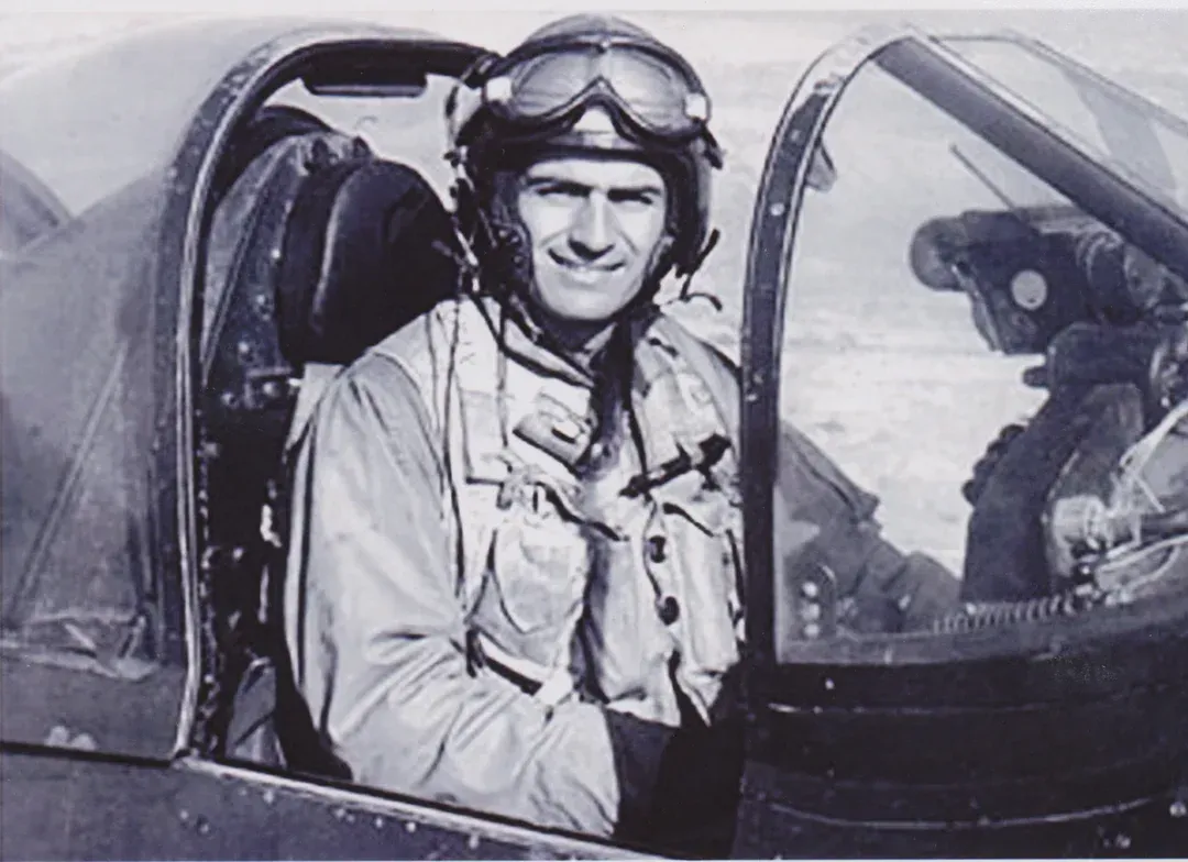 Pilot in cockpit wearing helmet and goggles, smiling.