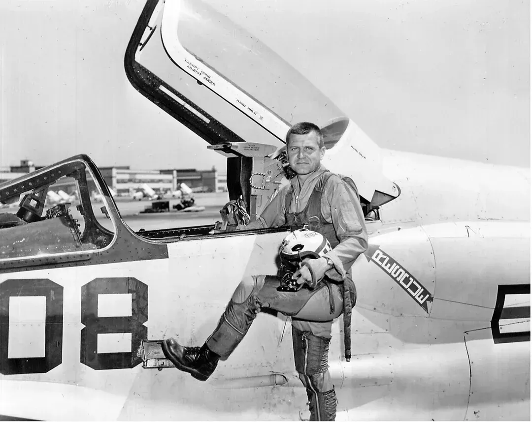 Pilot in flight suit next to a jet plane, stepping out of the cockpit, holding helmet, outdoors.