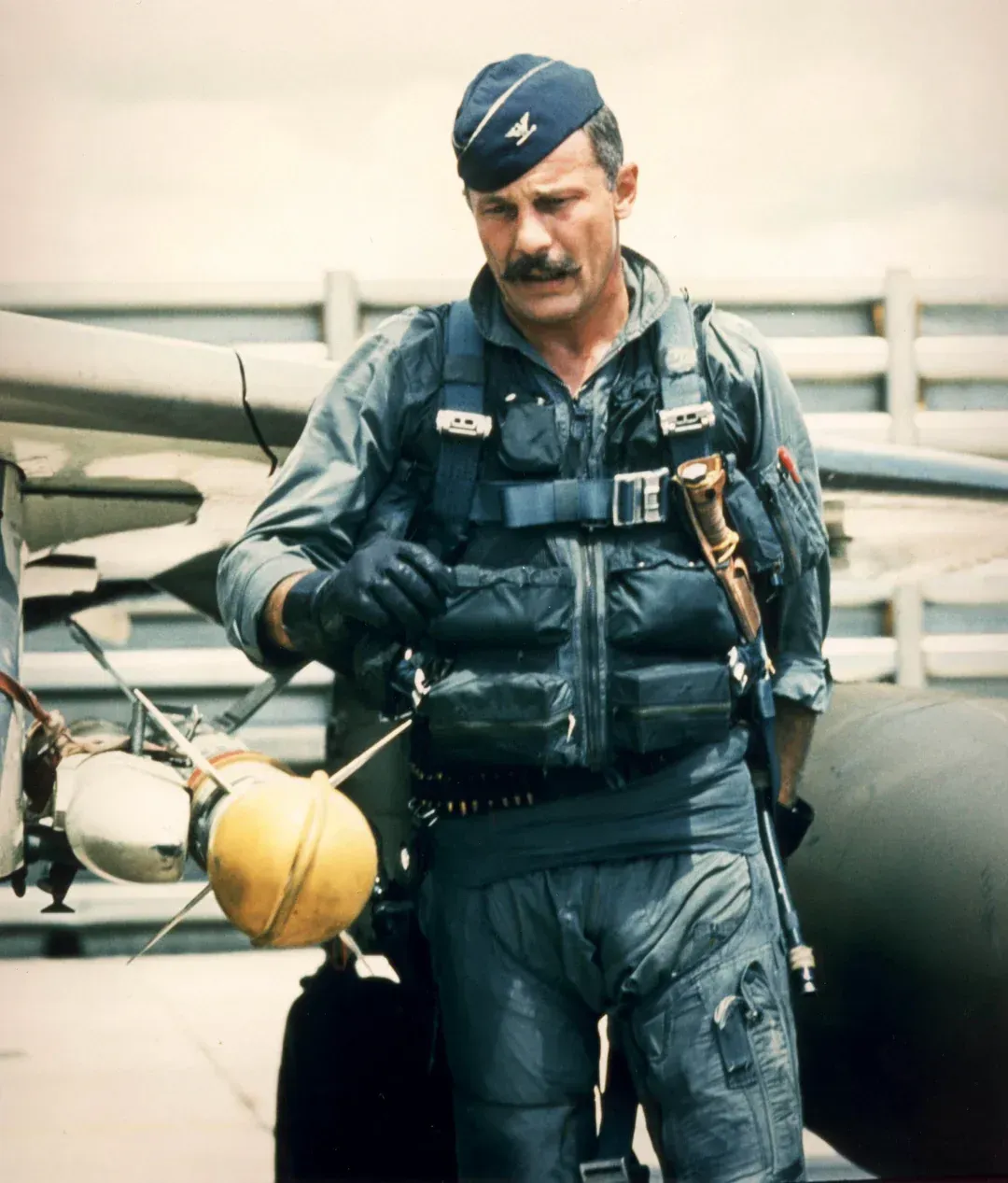 Pilot in blue flight suit, mustache, near jet. Wearing cap, holding glove. Outside, sunny.