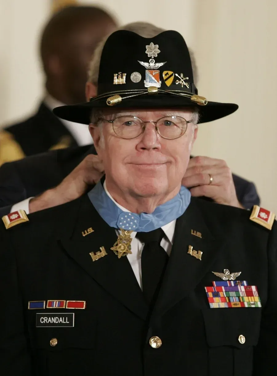Man in military uniform, receiving a medal at the White House. Smiling, wearing a hat adorned with pins.