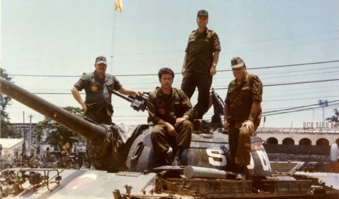 Soldiers on a tank, possibly during a parade, in an urban setting. Soldiers wear camouflage, a few are smiling.