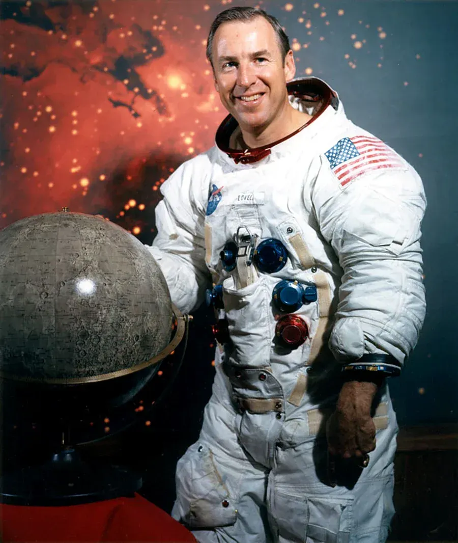 Astronaut smiling, in spacesuit, holding lunar globe, against an abstract red-orange background.