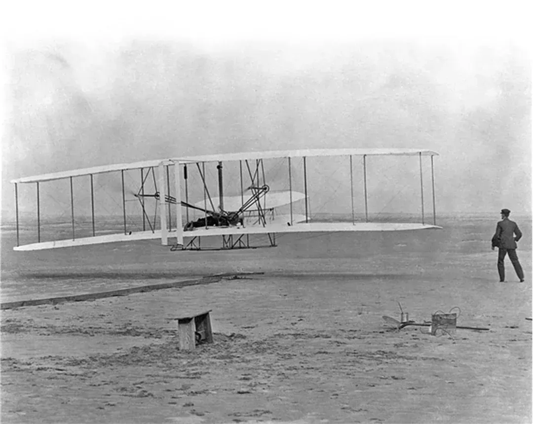 Wright brothers' first flight, a biplane with a pilot, launching on a sandy beach.