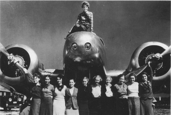 Group of women posing with a plane, one sits on the nose, propellors visible.