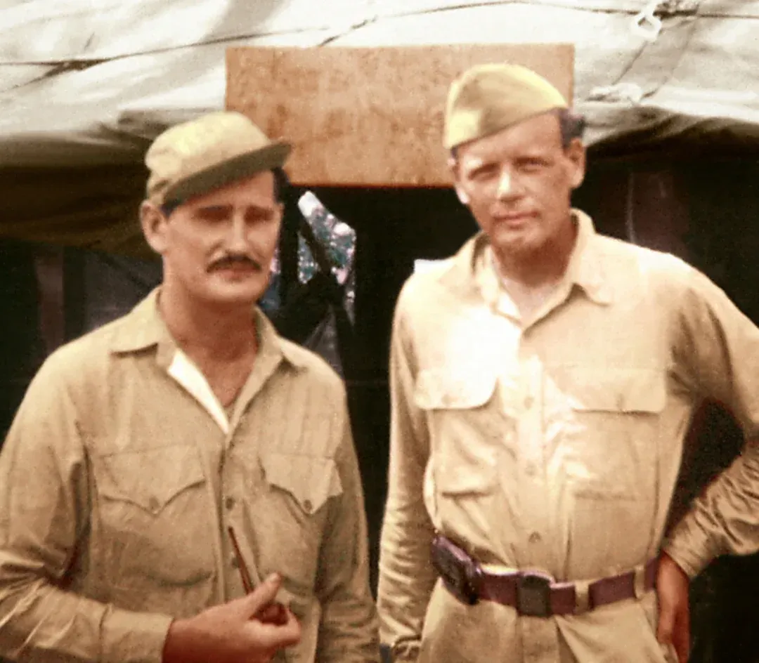 Two men in tan uniforms and caps stand outside a tent. One with a mustache.