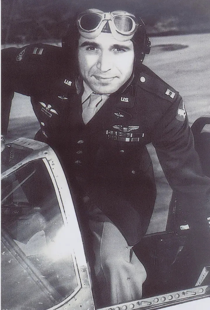 World War II pilot in uniform, leaning out of an airplane. He's wearing goggles and smiling.