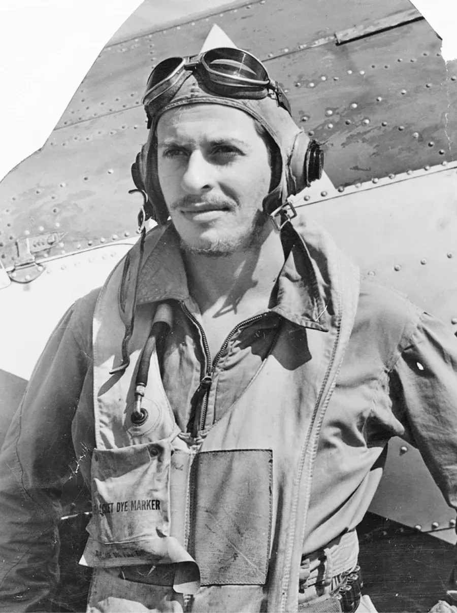 World War II pilot in uniform, goggles, and headset stands by a plane.