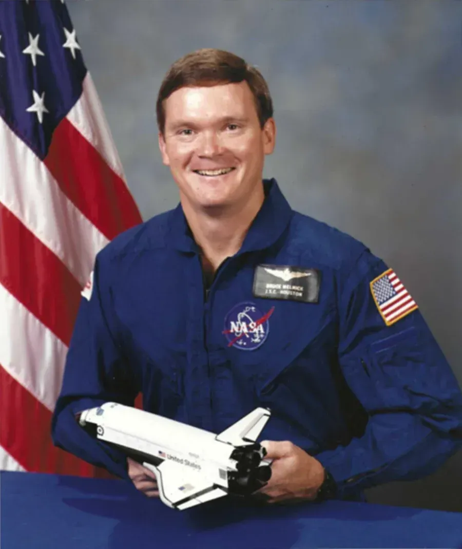 Astronaut in blue jumpsuit, holding a model space shuttle, smiling in front of the American flag.