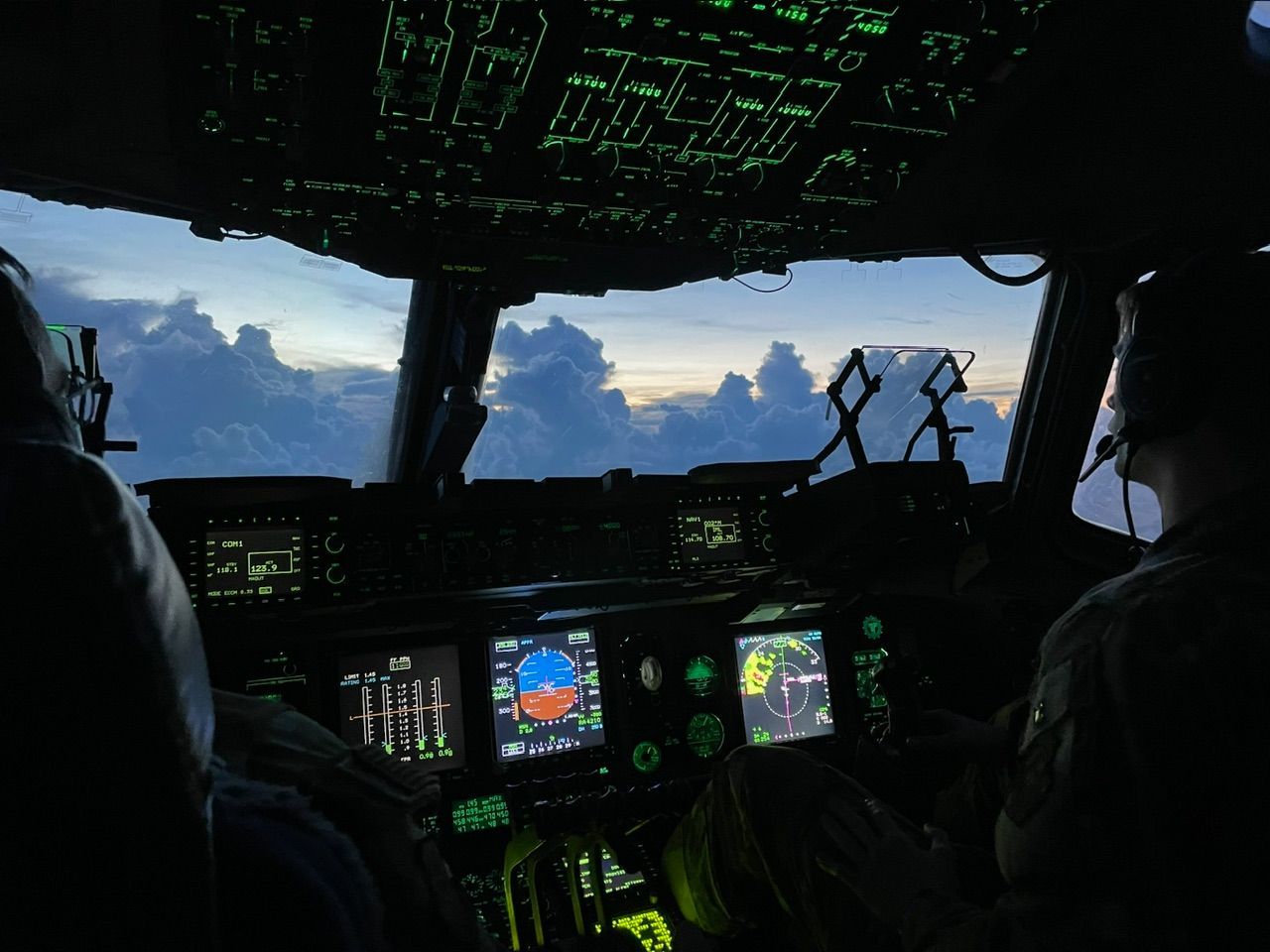 View from a cockpit at sunset. Pilots in silhouette against the sky, surrounded by illuminated control panels.