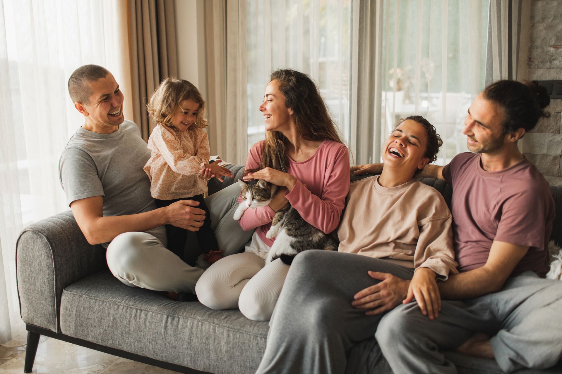 Family of five laughs together on a couch, petting a cat. Indoor, neutral colors.