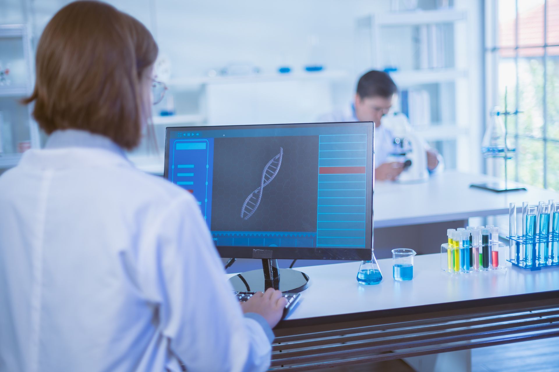 Scientist in lab coat looking at DNA helix on computer screen, another person in background.