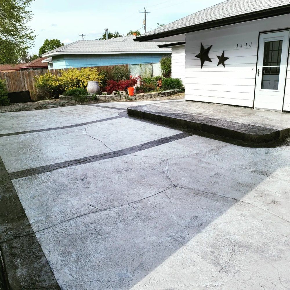 Concrete patio with gray cracked texture and dark accents, adjacent to a white house with stars and a garden.