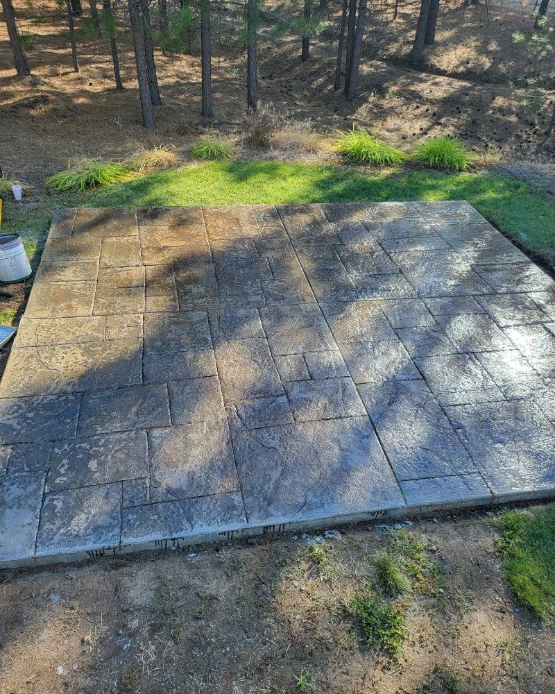 A gray brick patio with uneven surfaces, surrounded by grass and trees in a natural setting.