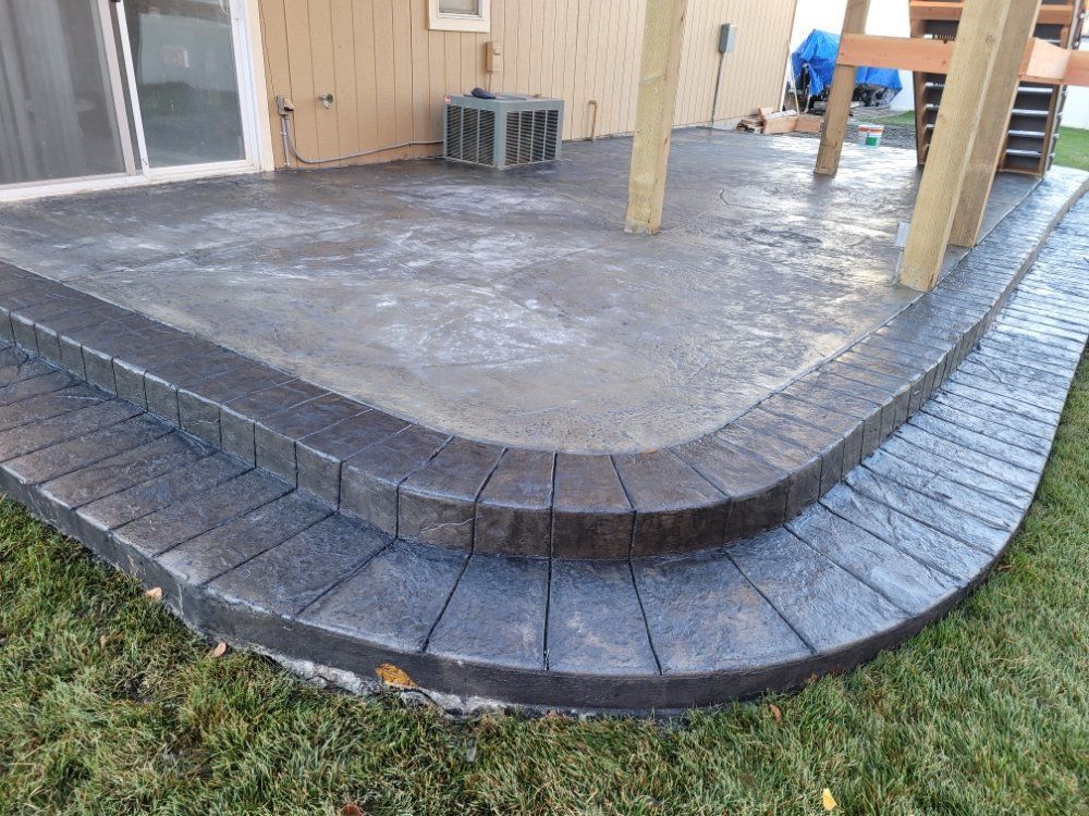 Concrete patio with curved steps, dark brick pattern border, next to grassy yard.