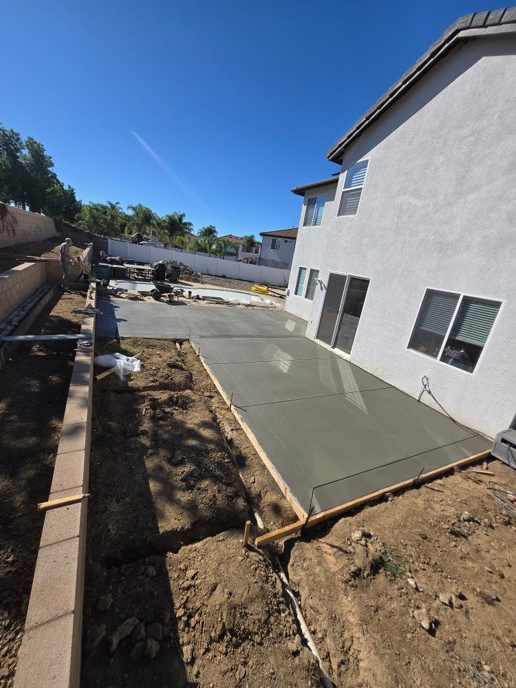 New concrete patio construction next to a house with a retaining wall on a sunny day.