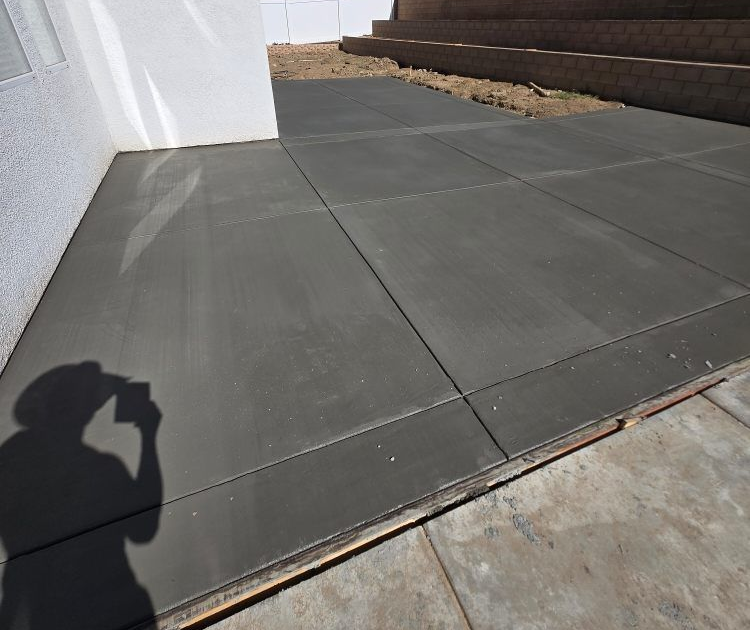 Newly poured gray concrete patio with dividing lines, next to a white wall and dirt.