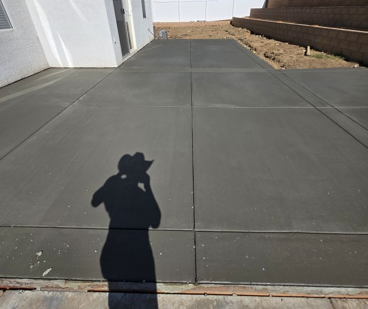 Newly poured concrete patio, gray, with expansion joints. Person's shadow visible. Sunny outdoor setting.