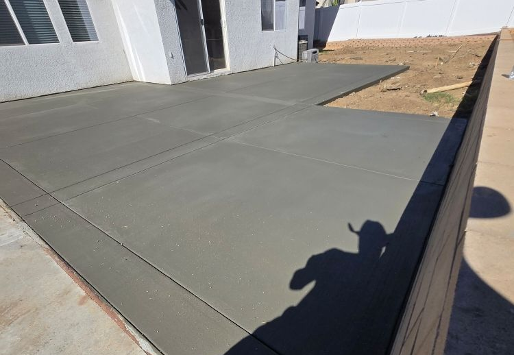 Newly poured concrete patio, gray, with expansion joints. Adjacent to a white building and brown wall.