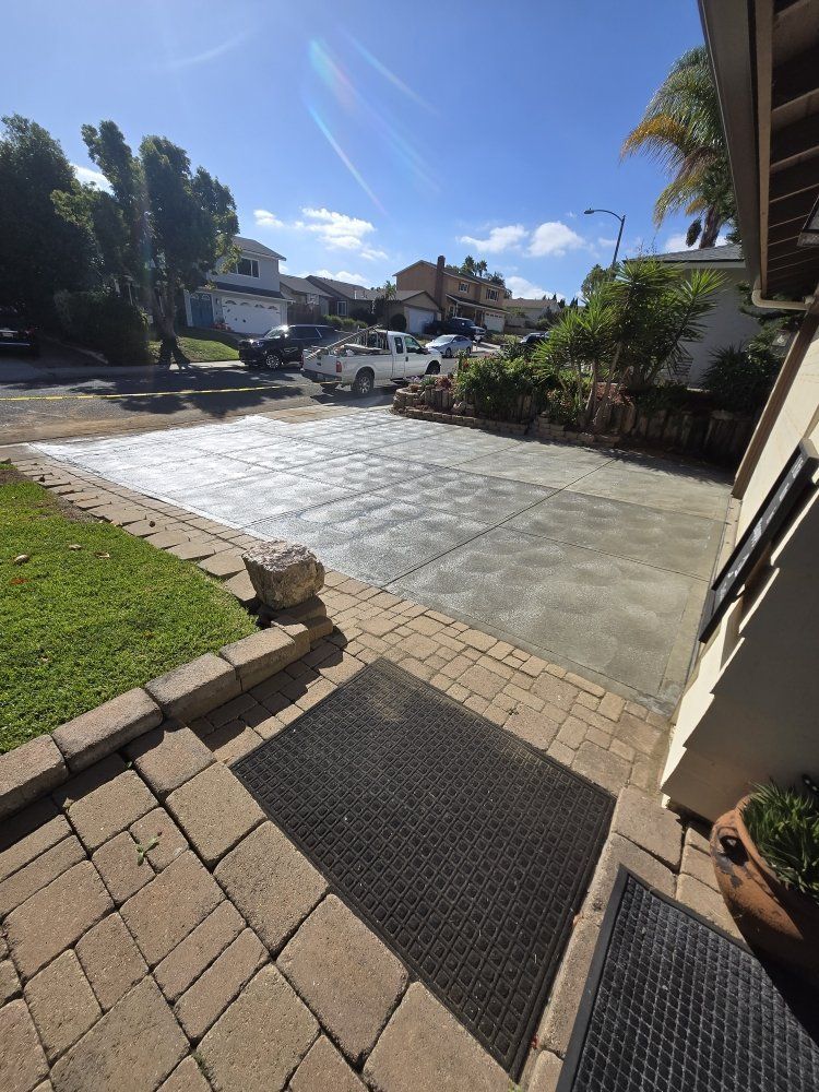 Driveway with a newly paved concrete section, lined by brick and a small lawn. Bright sun, blue sky.