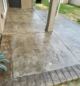 Concrete patio with stone border, column, and door. Wet with brown and gray tones, on lawn.