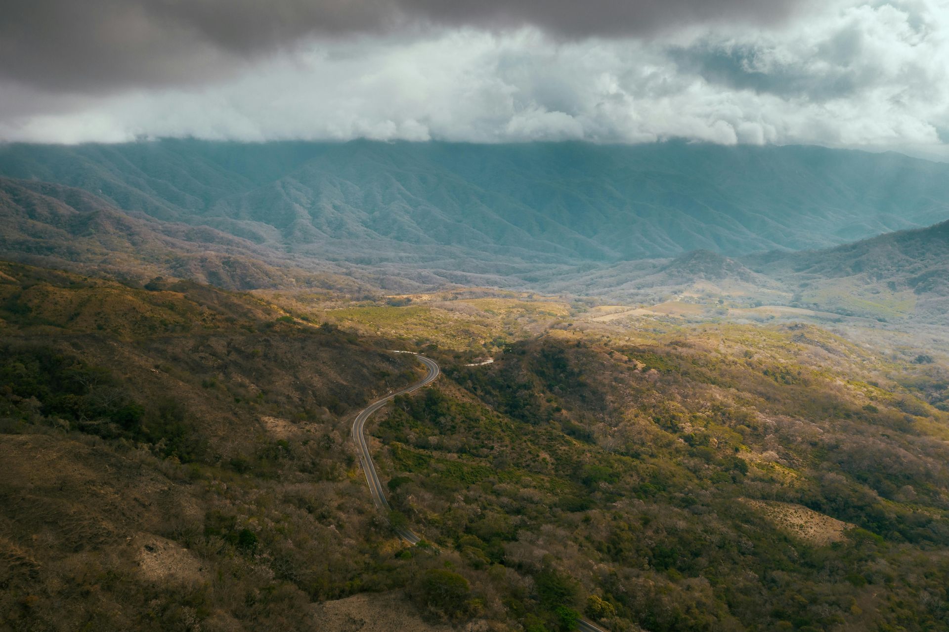 Rolling mountain landscape with a winding road through a valley, under cloudy skies.