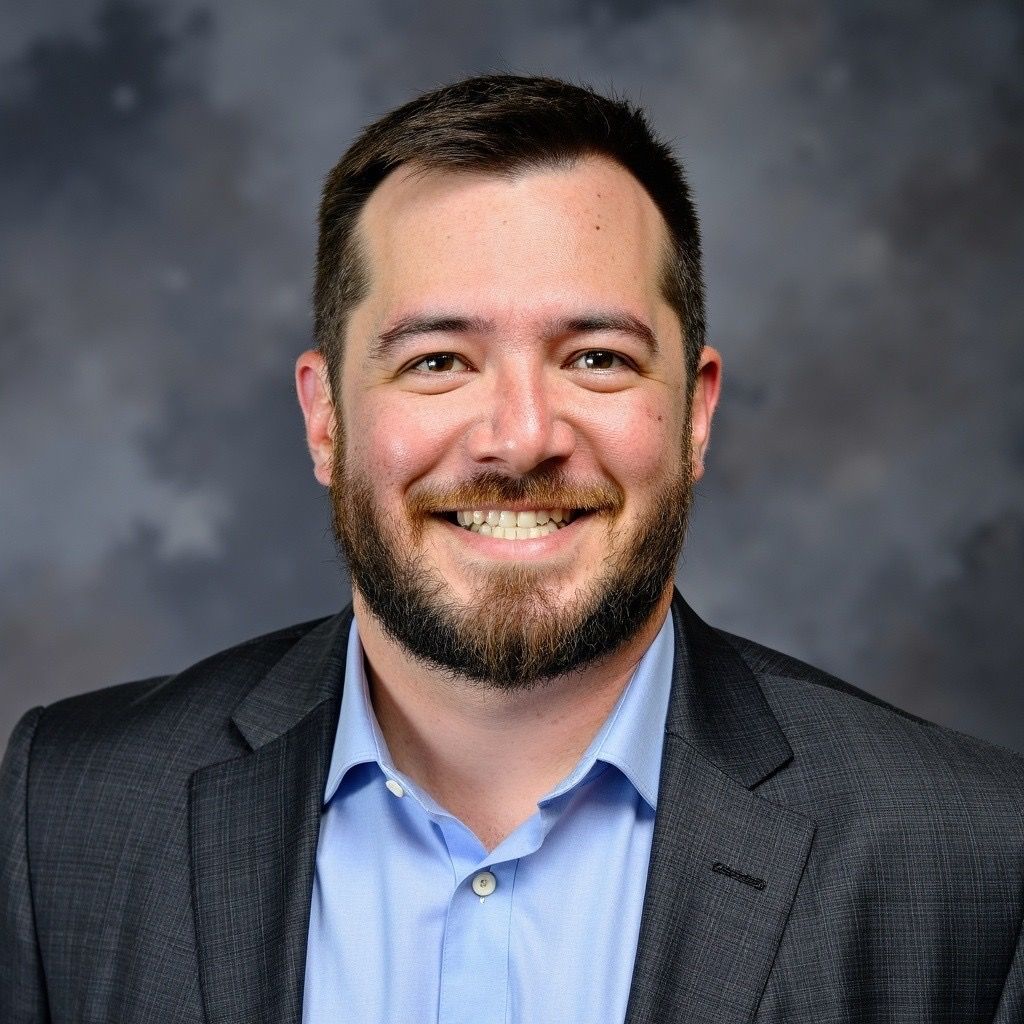 Man in a dark suit jacket, white shirt, smiling. Gray background.