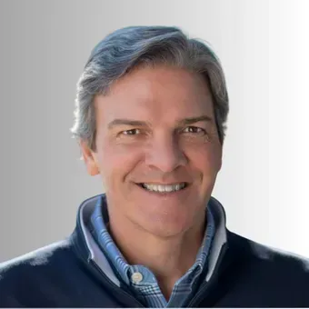 Man smiling, wearing a blue sweater over a collared shirt. Soft, blurred background.