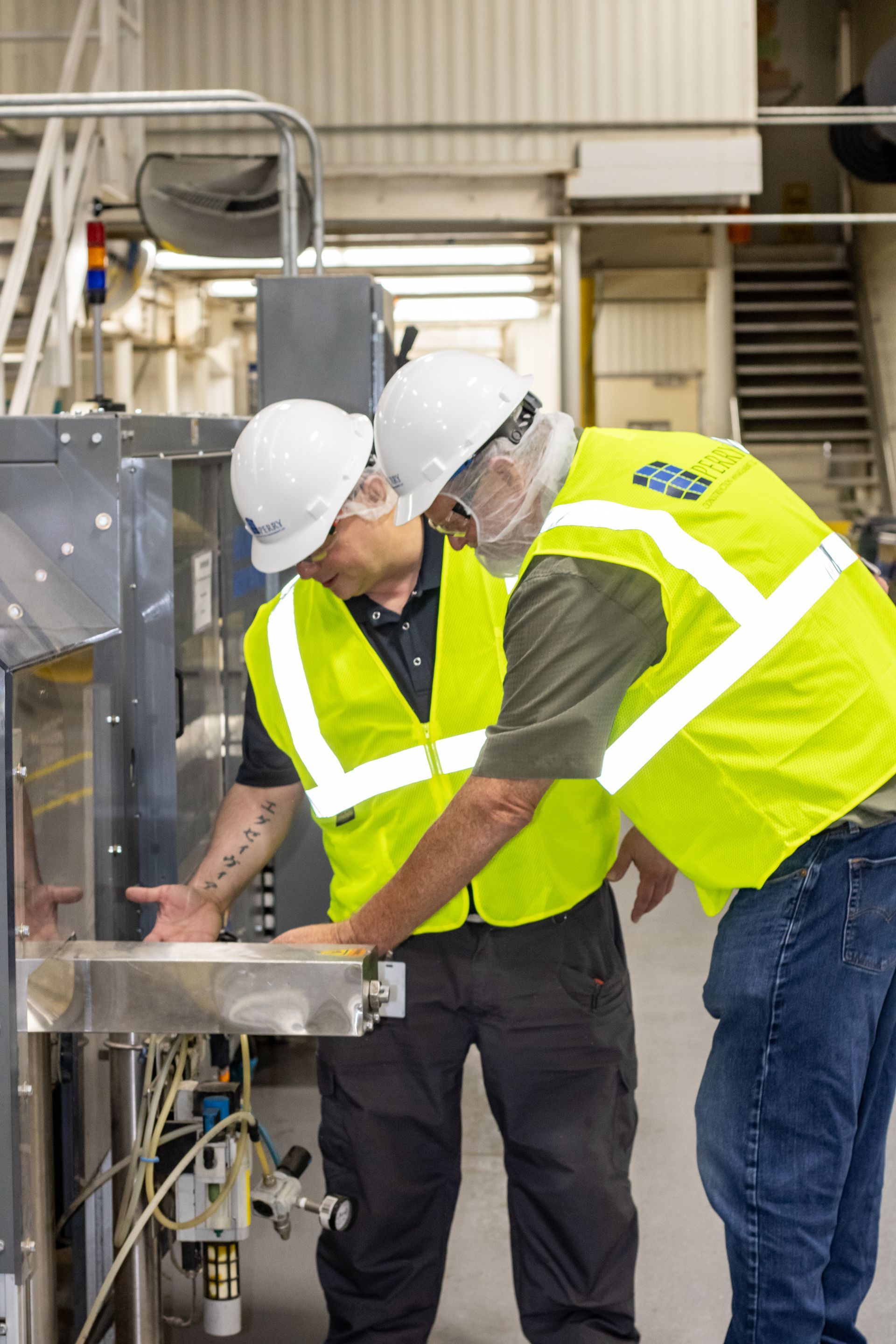 Two workers in safety vests and hard hats inspecting machinery in a factory.
