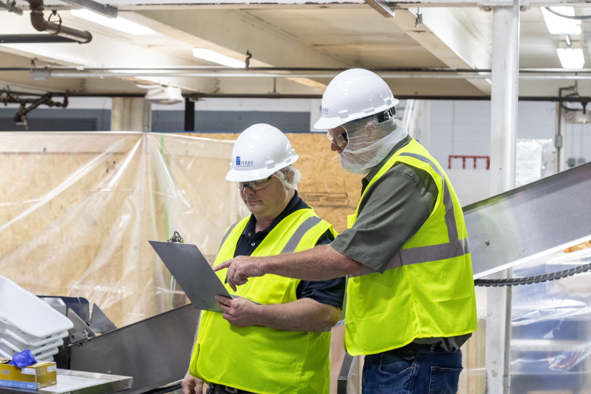 Two people in hard hats and vests review a document in a factory setting.