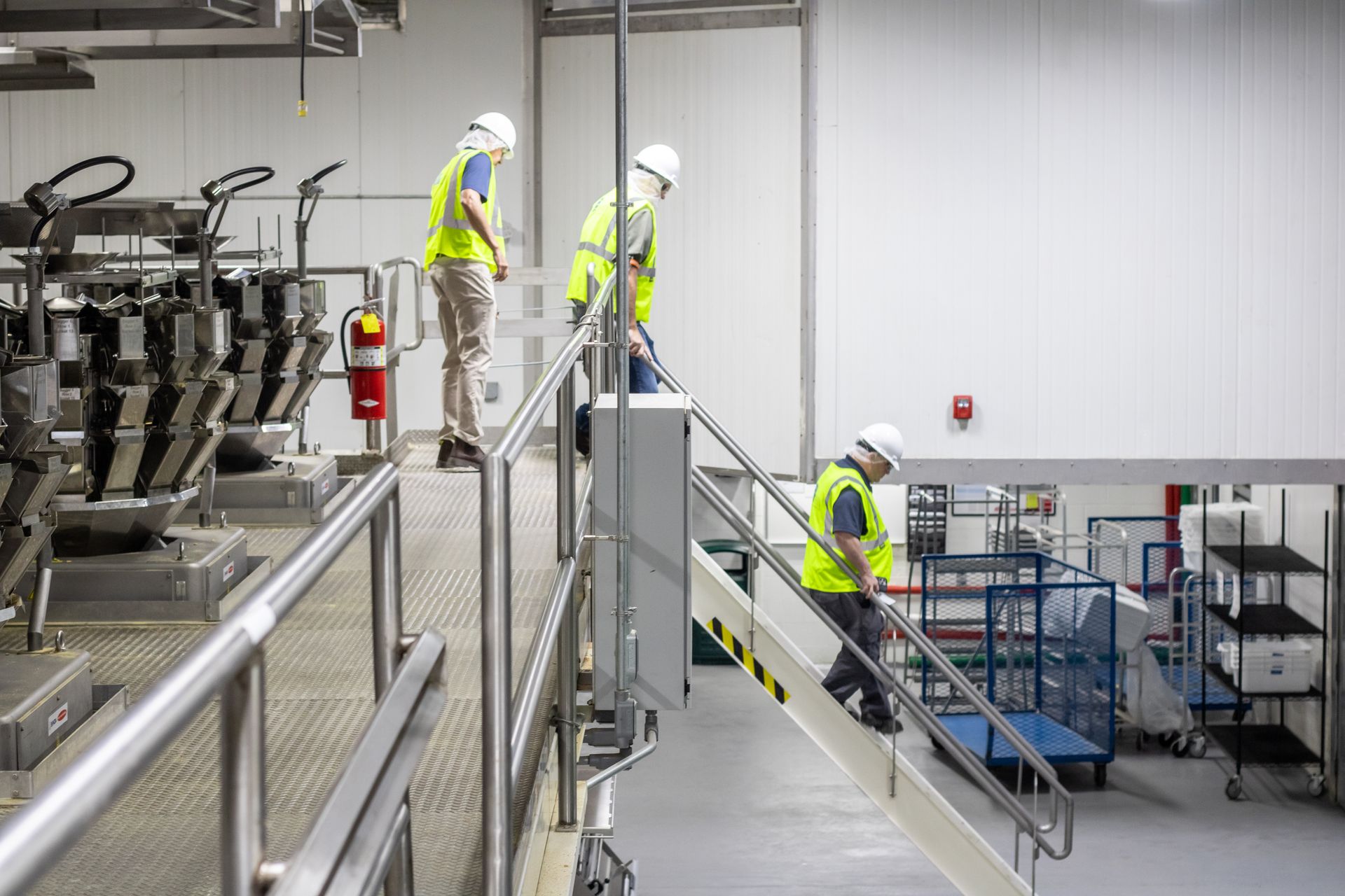 Three workers in safety vests and hard hats on a factory platform. One walks down stairs, two stand.