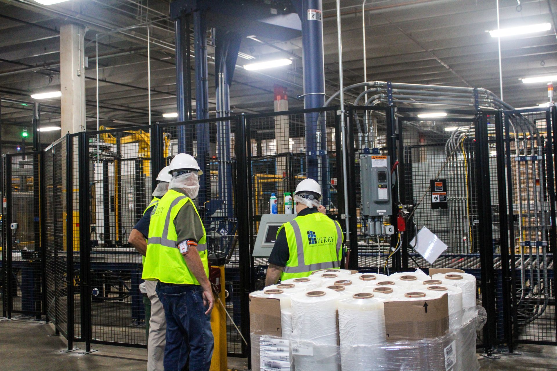 Workers in safety vests and hard hats operating machinery in a factory; rolls of paper products nearby.