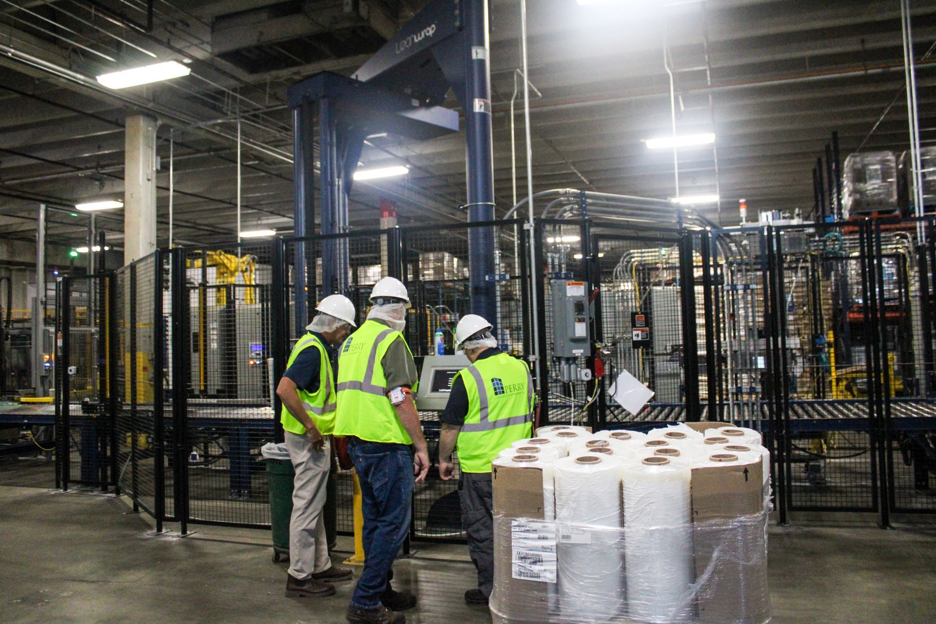 Three workers in safety vests examine machinery in a warehouse; rolls of wrapped paper in foreground.