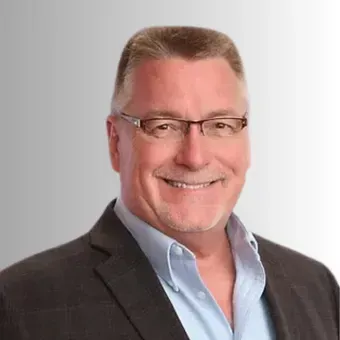 Man in glasses smiles, wearing a blazer over a blue button-down shirt, neutral background.