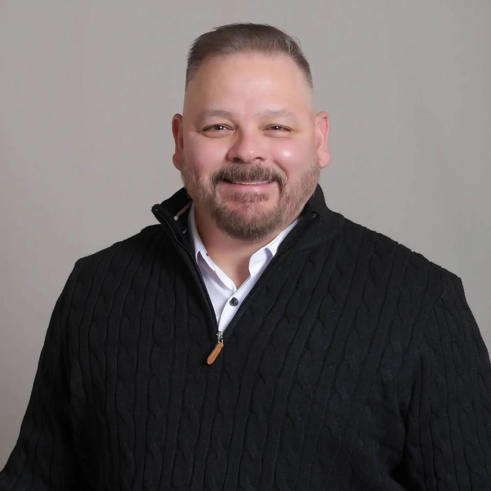Man with a neatly trimmed beard, wearing a black sweater over a white collared shirt, smiling in front of a gray backdrop.