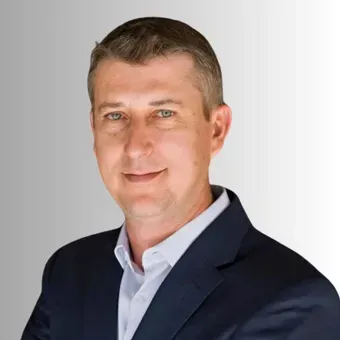 Man in a suit smiling, with a white shirt and dark blue jacket, against a gray gradient background.