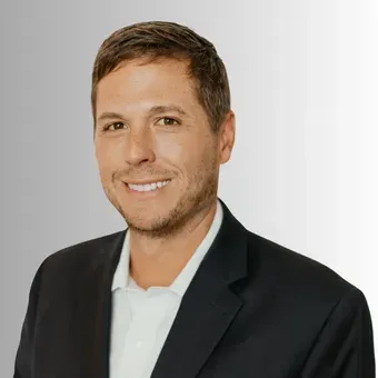 Man in a dark suit jacket, white shirt, smiling. Gray background.