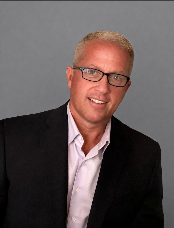 A person wearing glasses and a dark blazer over a light shirt smiles against a plain gray background.