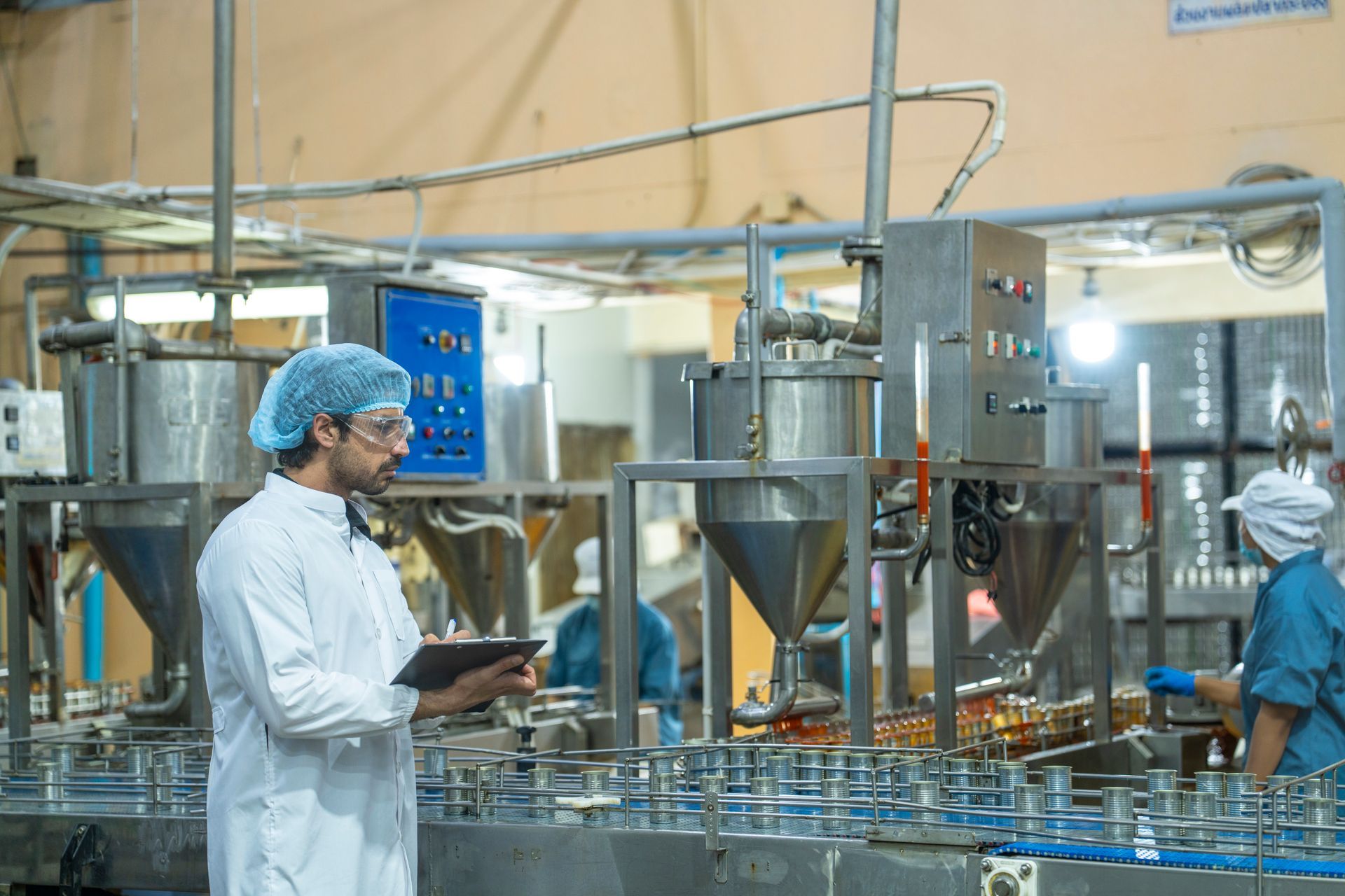 Man in lab coat inspects factory equipment, holding tablet. A worker operates machinery. Industrial setting.