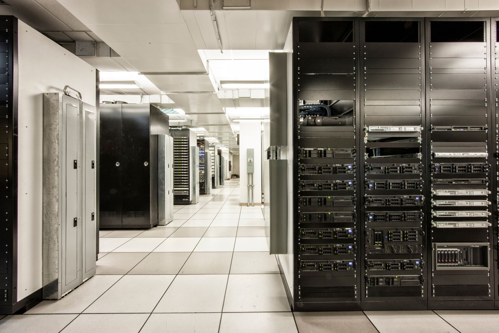 Rows of server racks in a data center, lit by overhead lights, tile floor.