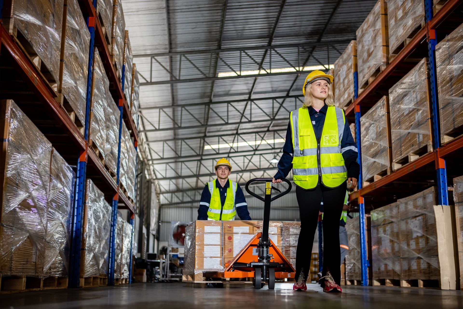 Two workers in a warehouse wearing safety vests and hard hats, one operating a pallet jack.