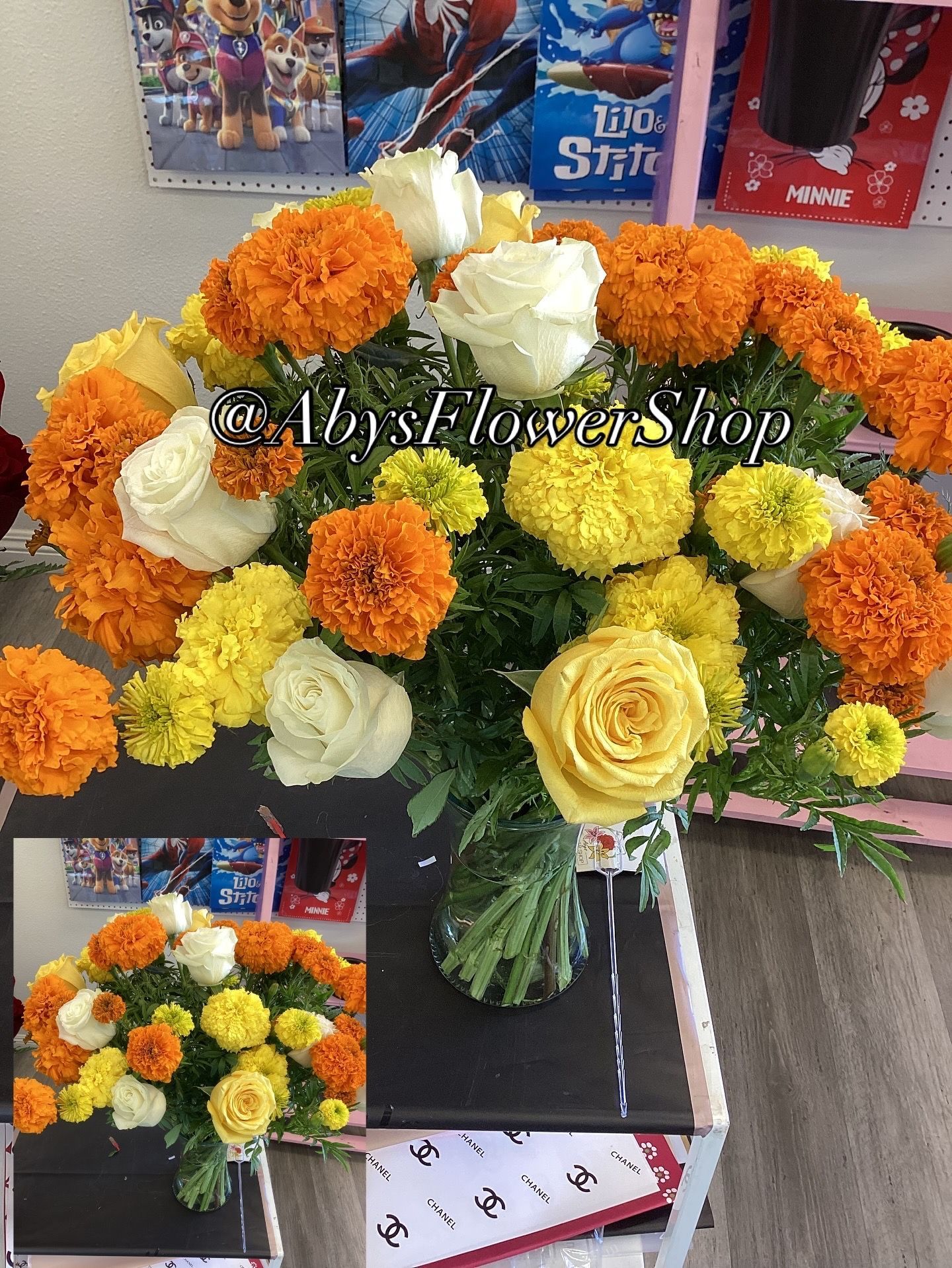 A vibrant bouquet of orange and yellow marigolds mixed with white and yellow roses in a glass vase on a display table.