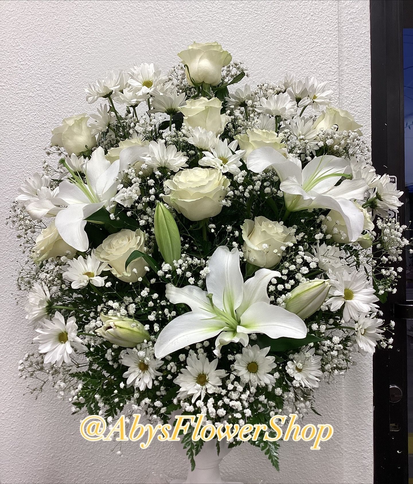 A large, rounded floral arrangement of white roses, lilies, and daisies with baby's breath, set against a plain background.