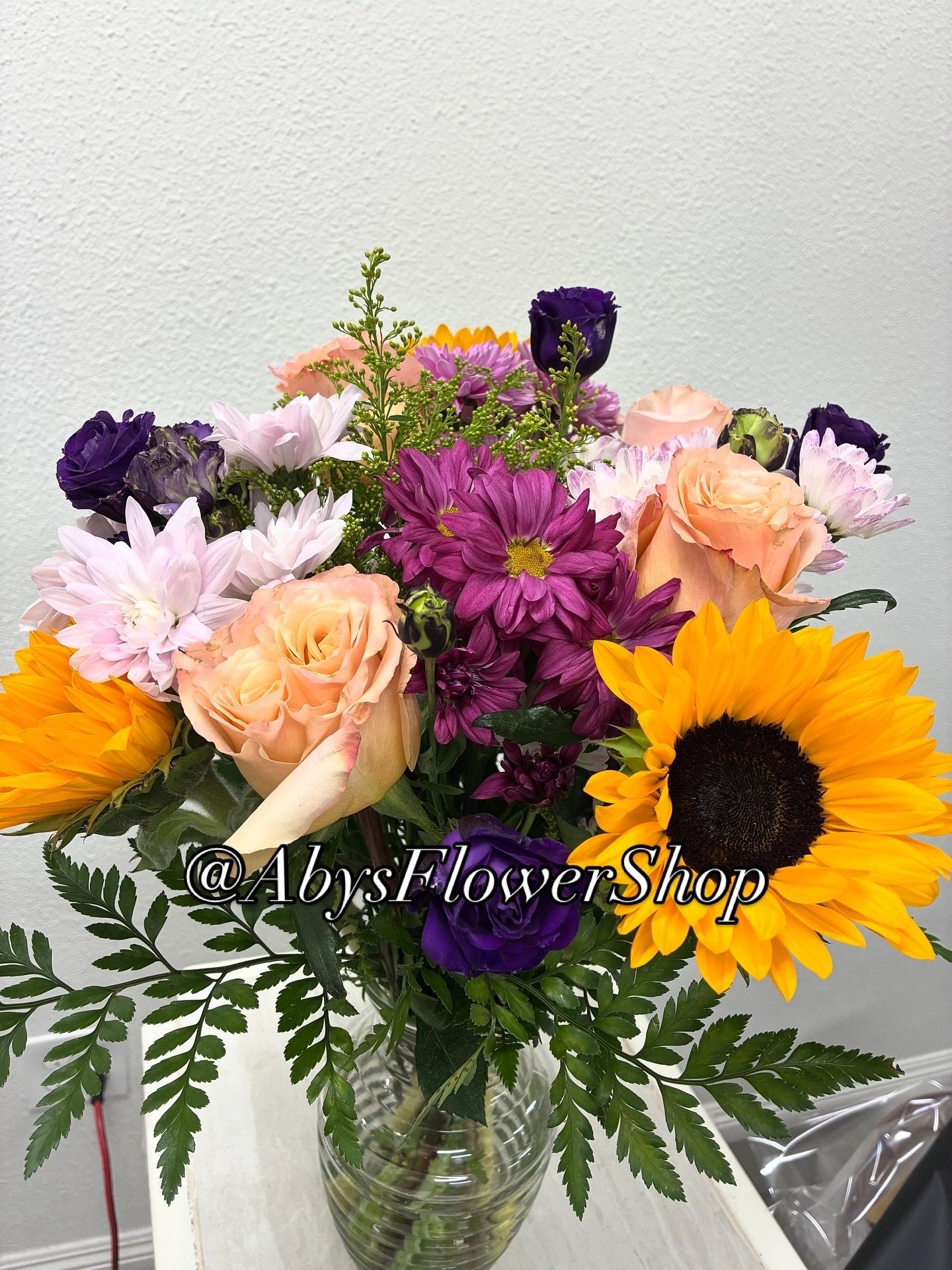 A bouquet featuring sunflowers, peach roses, and purple chrysanthemums in a glass vase, with a floral shop watermark.