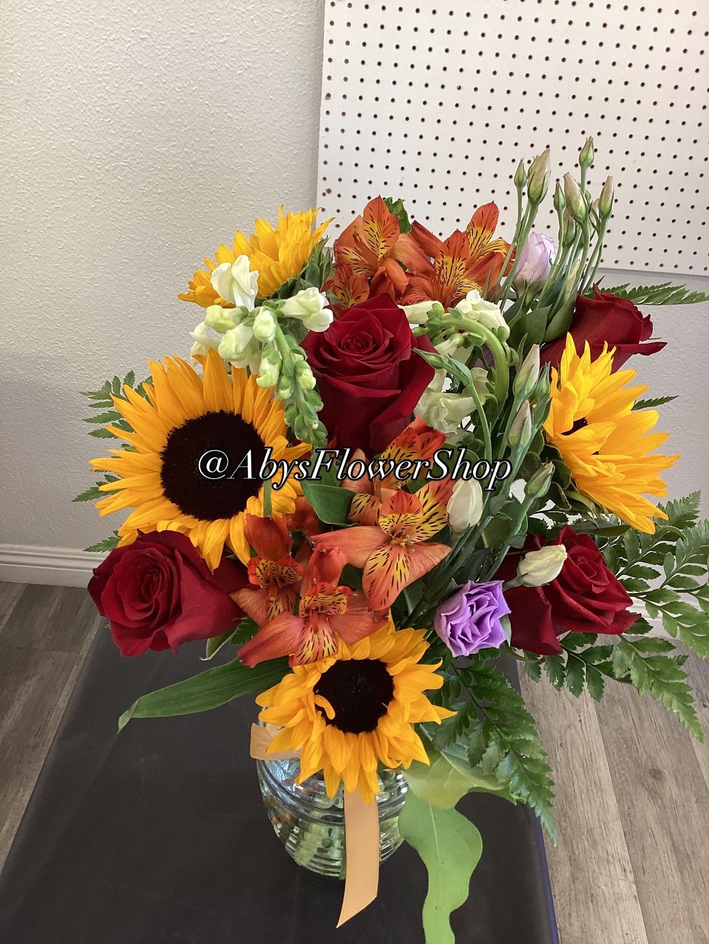 A vibrant floral bouquet featuring sunflowers, deep red roses, orange alstroemeria, and greenery in a clear glass vase.