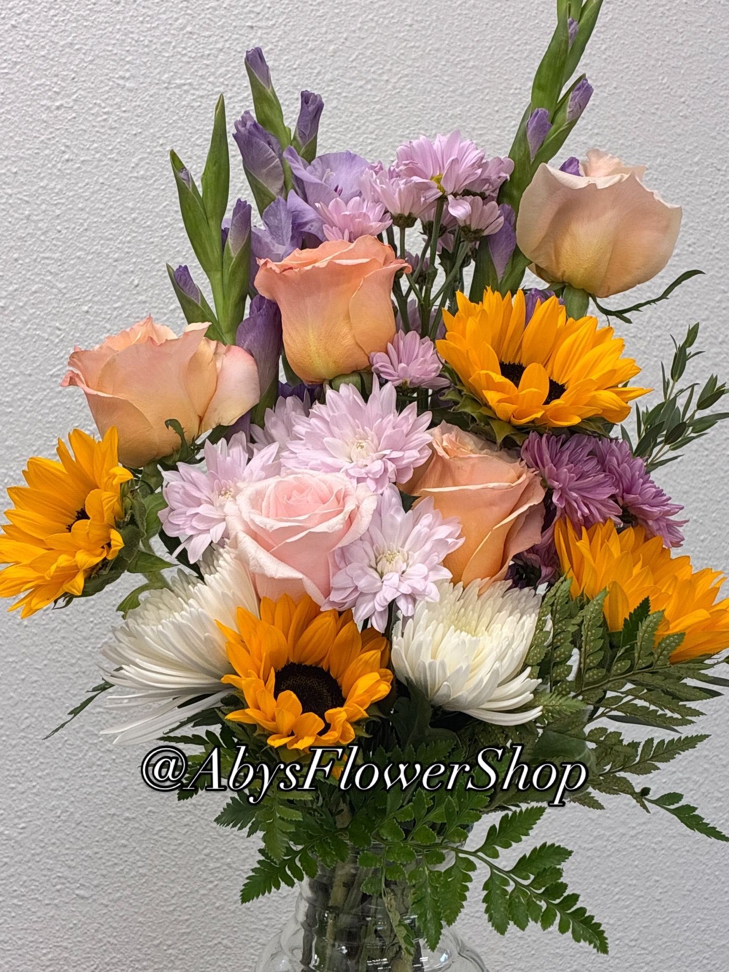 A bouquet with yellow sunflowers, soft orange roses, purple gladiolus and mums, and green fern foliage in a vase.