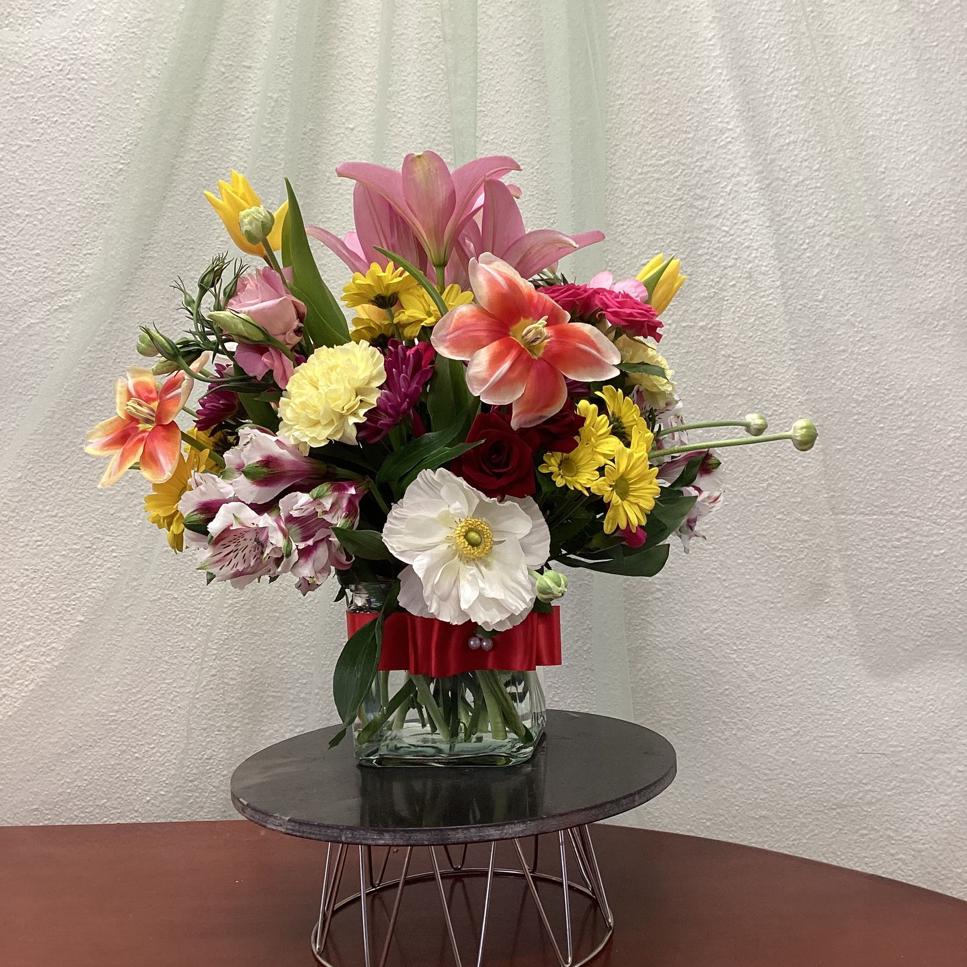 A colorful floral arrangement in a glass vase with a red ribbon, displayed on a circular black stand against a white backdrop.