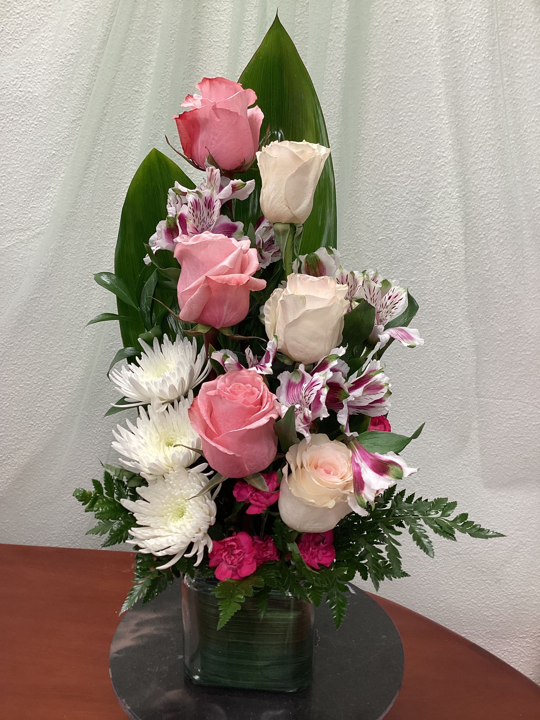 A floral arrangement featuring pink and white roses, white chrysanthemums, and purple-speckled lilies in a glass vase.