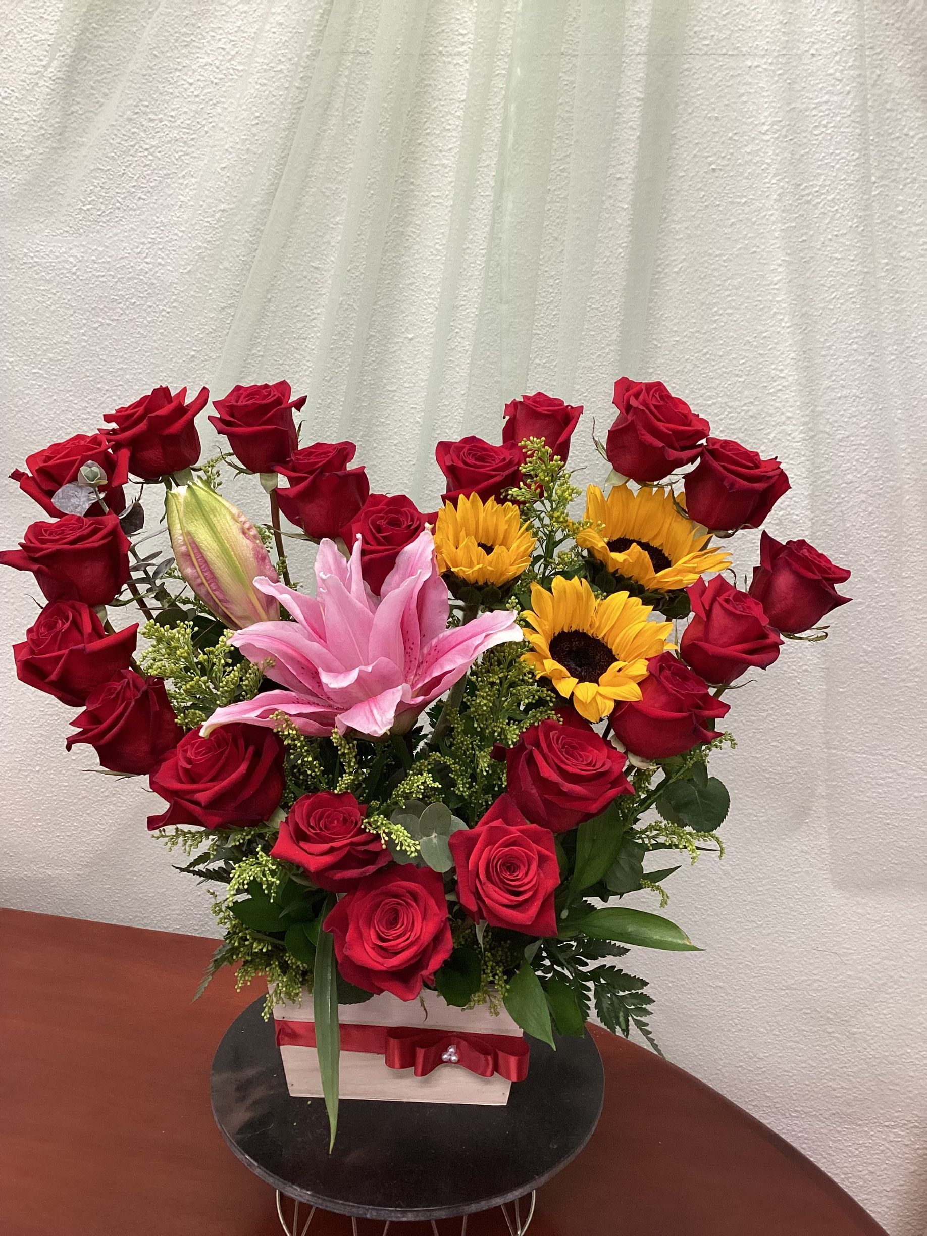 A heart-shaped floral arrangement with red roses, yellow sunflowers, and a large pink lily in a box on a stand.