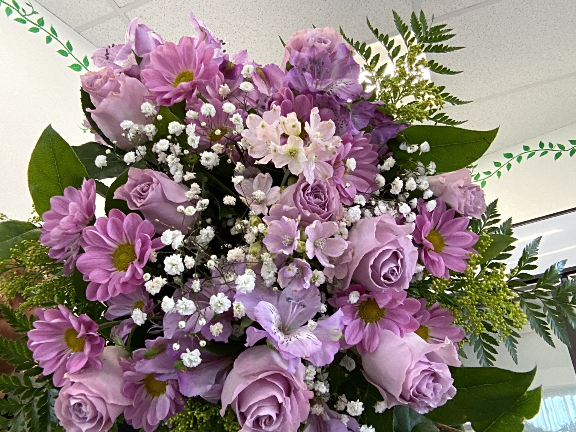 A lush floral bouquet featuring lavender roses, purple daisies, and white baby's breath with green foliage.