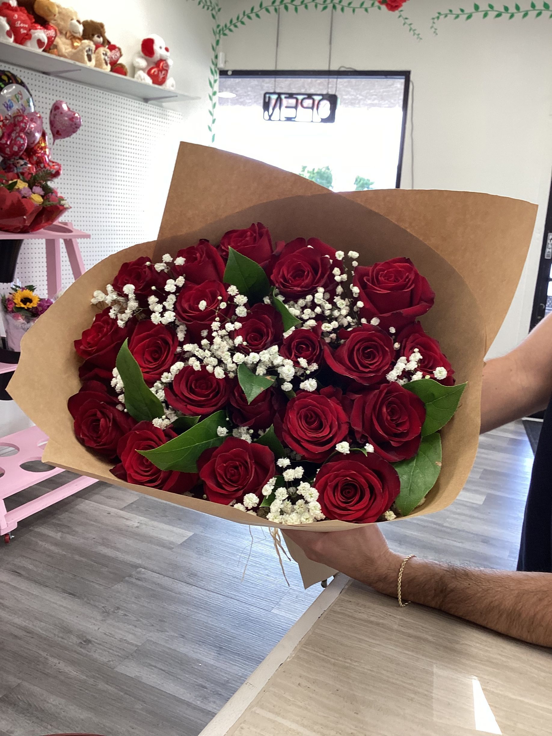 A hand holds a bouquet of red roses accented with white baby's breath, wrapped in brown paper inside a floral shop.
